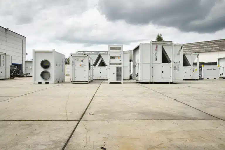 Een buitenterrein met diverse industriële koelinstallaties en luchtbehandelingsunits van Delta-Temp, opgesteld op een betonnen ondergrond. De apparaten zijn wit en voorzien van het Delta-Temp-logo. De achtergrond toont een industrieel gebouw en een bewolkte lucht.
