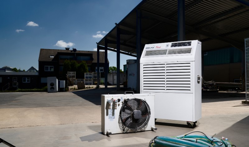 Twee witte Delta-Temp airconditioningunits, één rechtopstaand en één liggend met een ventilator, staan buiten op een betonnen oppervlak onder een afdak op een zonnige dag.