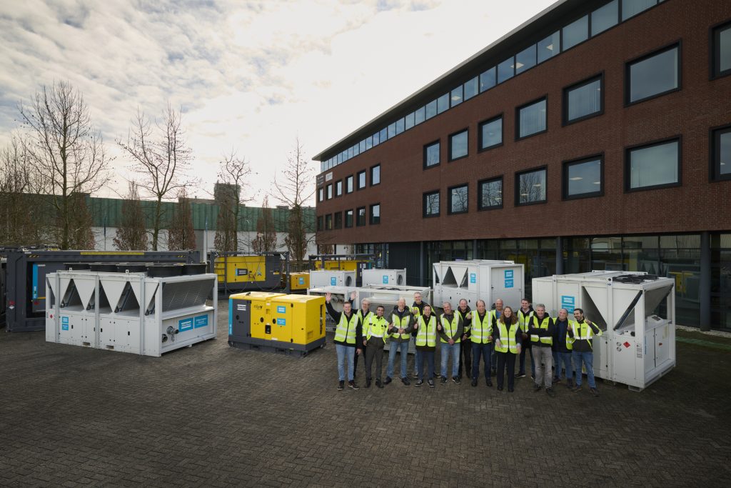 Een groep medewerkers in reflecterende veiligheidsvesten die poseren voor een collectie chillers, airhandlers en generatoren bij een kantoorpand.